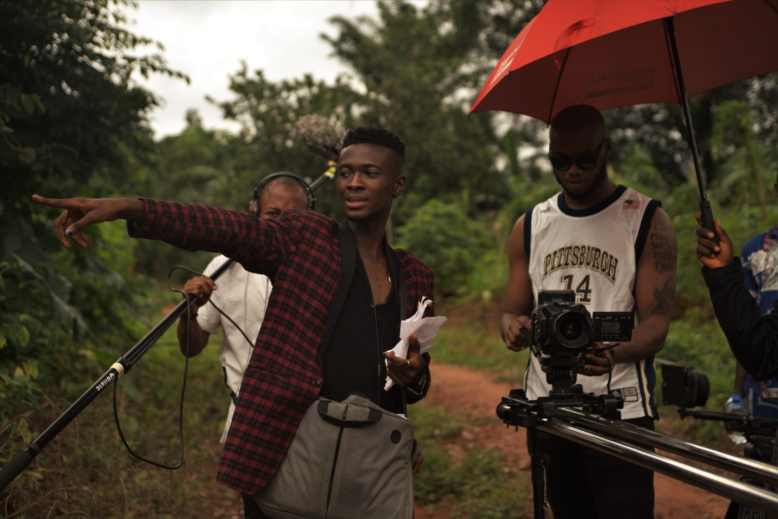 Image description: Wapah Ezeigwe on set of Country Love. The image shows Wapah Ezeigwe directing a scene from Country Love. They are dressed in a black shirt and a red and black striped jacket with a grey bag hanging across their shoulders. They are pointing at something with one hand and holding a bunch of papers in another. Standing by them are three persons. One of them dressed in a white basketball vest is holding a camera and the other dressed in a white T-shirt too is holding a boom microphone. There is a visible hand holding up a red umbrella for the rest. 