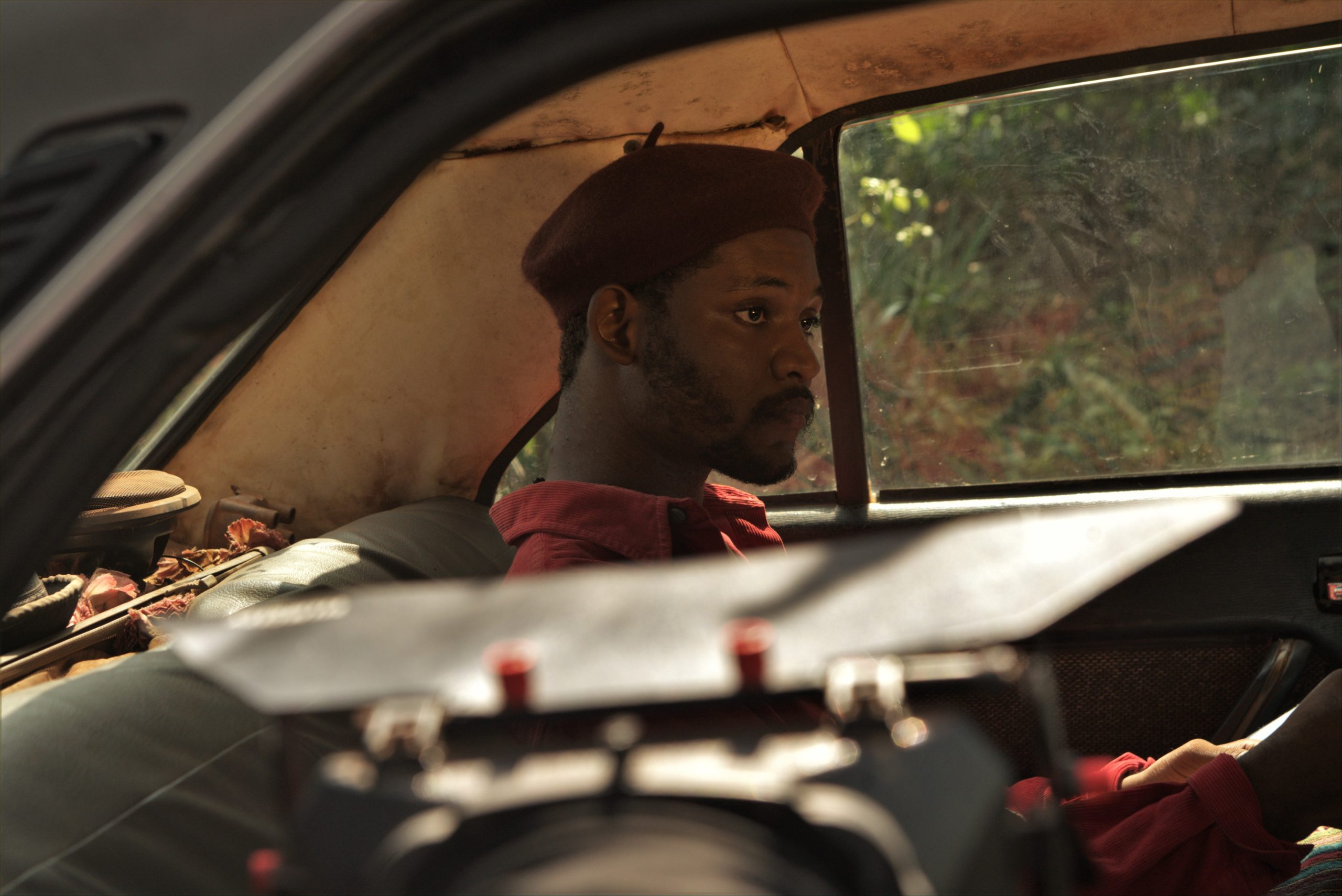 Image description: Kelechi Michaels in the film Country Love. The character, Kambili is sitting in the backseat of a car. There is a trunk beside him. He is dressed in an orange shirt and wearing a maroon beret. 